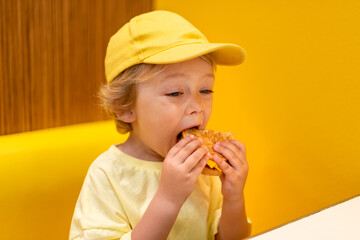 Young child wearing a yellow hat and shirt eats a cheeseburger in a vibrant restaurant, enjoying fast food meal in a casual dining environment. Kids diet, bad food choices, early junk food addiction