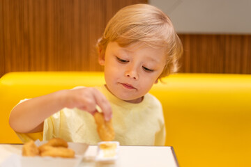 A small child wearing a yellow shirt is eating chicken nuggets in a vibrant environment. Four year old boy enjoying fast food meal. Kids diet, bad food choices, early junk food addiction