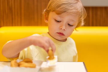 A small child wearing a yellow shirt is eating chicken nuggets in a vibrant environment. Four year old boy enjoying fast food meal. Kids diet, bad food choices, early junk food addiction