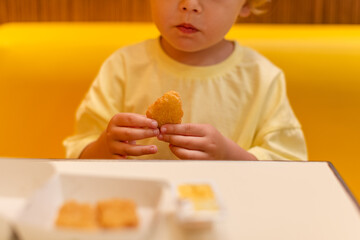 A small child wearing a yellow shirt is eating chicken nuggets in a vibrant environment. Four year old boy enjoying fast food meal. Kids diet, bad food choices, early junk food addiction