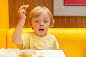 A small child wearing a yellow shirt is eating chicken nuggets in a vibrant environment. Four year old boy enjoying fast food meal. Kids diet, bad food choices, early junk food addiction