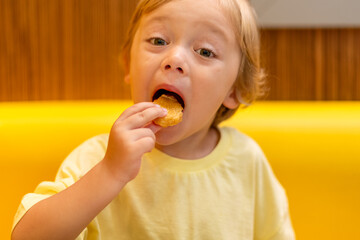 A small child wearing a yellow shirt is eating chicken nuggets in a vibrant environment. Four year old boy enjoying fast food meal. Kids diet, bad food choices, early junk food addiction