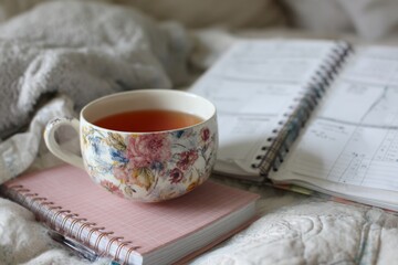 Floral teacup sits atop a pink notebook, calendar journal is slightly blurred in soft morning light on cozy blanket.