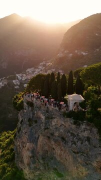 Aerial view of luxury wedding at Villa Cimbrone, Ravello. Guests dance on a cliff-edge terrace above a sheer stone wall, surrounded by red umbrellas, sea haze, and golden Amalfi Coast sunset