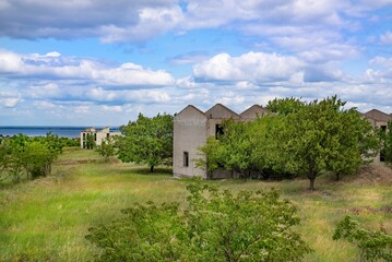 Row of derelict concrete buildings surrounded by wild vegetation near a coastline under a cloudy...