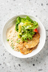 A bowl of creamy risotto with peas, accompanied by a fresh green salad with lettuce and two crispy fried fish fillets, presented on a speckled gray surface. A balanced and appetizing meal with vibrant
