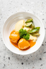 A plate of mashed potatoes with two pieces of meat drizzled in orange sauce, garnished with fresh parsley and accompanied by green vegetables, served on a white bowl against a speckled countertop