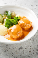 A plate of mashed potatoes with two pieces of meat drizzled in orange sauce, garnished with fresh parsley and accompanied by green vegetables, served on a white bowl against a speckled countertop