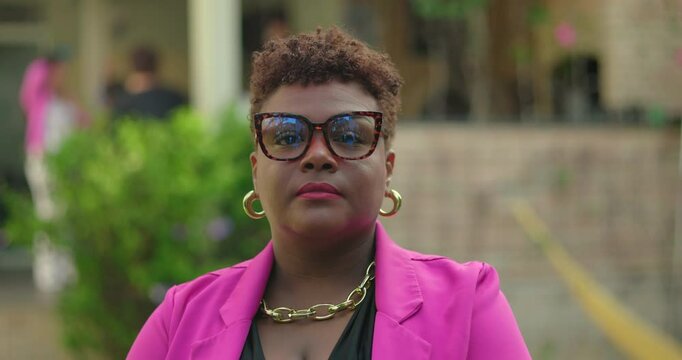 Portrait of African American woman in pink blazer and glasses standing outdoors, looking forward with assertive presence and modern fashion style