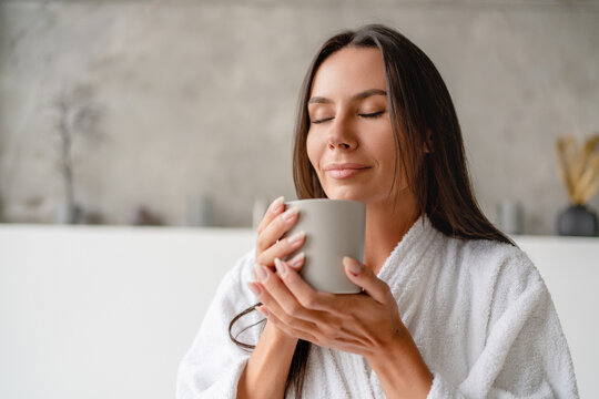 Good morning. Relaxed brunette woman drinking coffee holding cup sitting on bathtub wearing white bathrobe in luxury bathroom indoor. Pampering and beauty rituals concept