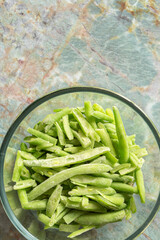 Frozen sliced green beans defrosting in a glass bowl background backdrop. On a green marble background.