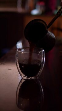 An intimate and authentic mood is captured as rich, dark Turkish coffee is poured from a traditional cezve into a modern double-walled glass, with subtle reflections on the surface