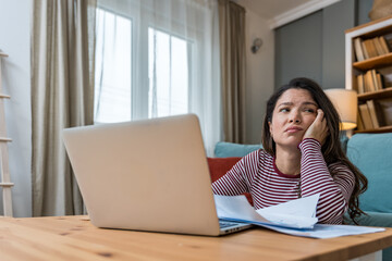 Young woman struggling with focus while working on laptop on floor, surrounded by printed analytic,...