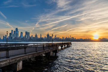 Pier in Hoboken, NJ extending over the Hudson River with New York City skyline in the background, bathed in early morning golden light under a dramatic sky.