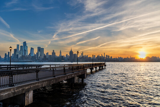 Pier in Hoboken, NJ extending over the Hudson River with New York City skyline in the background, bathed in early morning golden light under a dramatic sky.