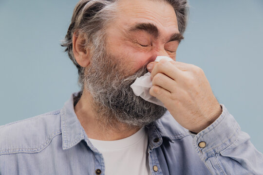 Senior man with gray beard and denim shirt blowing his nose with tissue