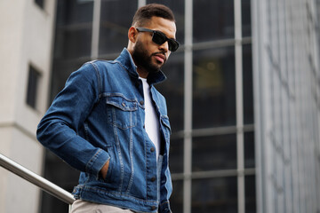 Stylish black man in sunglasses holding hands in pockets of denim jacket on street