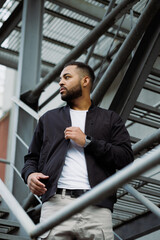 African american man wearing bomber jacket standing on fire escape on street
