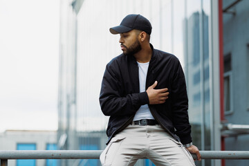 Stylish african american man in baseball cap sitting on railing on urban street