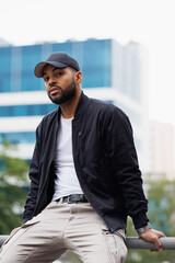 Young african american man in bomber jacket sitting on railing on city street