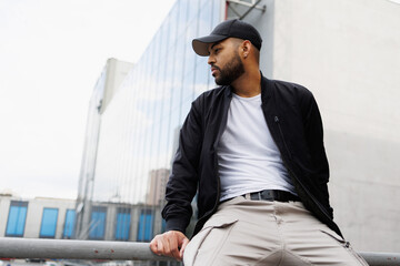 African american man in baseball cap and bomber jacket sitting on railing on urban street