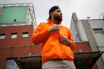 African american man in windbreaker and baseball cap standing near building on street