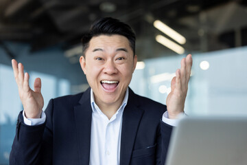 Portrait of a young Asian businessman sitting at a desk with a laptop and rejoicing in success