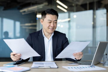 Asian young male businessman sitting in the office at a desk in front of a laptop, holding and working with documents