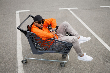 Trendy african american man in sunglasses and windbreaker jacket sitting in shopping cart on street