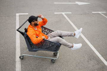 Positive african american man in street wear and sunglasses sitting in shopping cart on parking