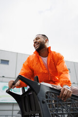 Laughing african american man in windbreaker sitting in shopping cart on street