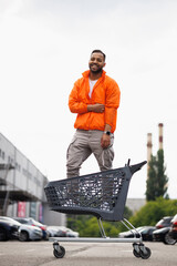 Cheerful african american man in windbreaker jacket standing in shopping cart on street