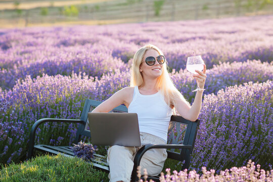 Lavender field. Woman sitting on bench among lavender flowers and working on laptop computer with glass of summer refreshing cocktail, relaxing, resting, enjoying nature. Mental health concept