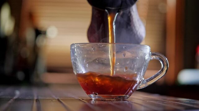 An inviting and comforting mood is captured as a stream of hot, dark tea or coffee is poured from above into a clear glass cup, highlighting the beverage's rich color and warmth