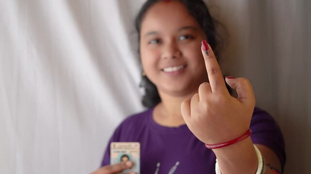 Selective focus on ink-marked finger of an Indian village woman with smiling face and voter card on other hand.