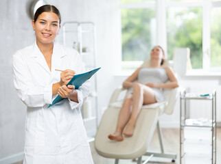 Obraz premium Young woman cosmetologist in uniform makes notes in medical room