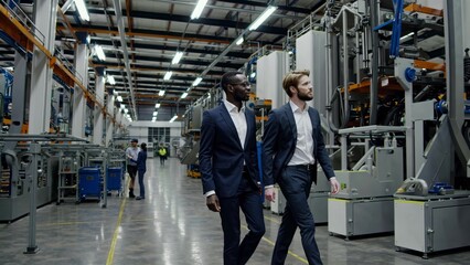 Two managers are having a business meeting while walking through a large, modern industrial factory, discussing production and manufacturing processes