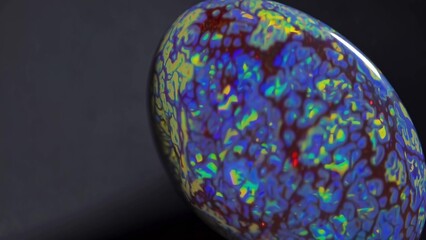 Polished opal gemstone displays a mesmerizing play of color, with iridescent blues, greens, and reds swirling across its surface against a dark background, creating a captivating visual spectacle