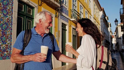 Senior man and woman tourists are enjoying a sunny day, holding disposable coffee cups and laughing together on a typical lisbon street in portugal