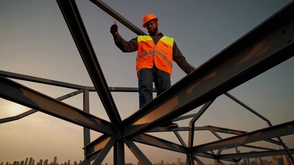 Confident african american construction worker showing thumbs up while carefully walking on steel beams of building under construction at sunset, symbolizing successful completion of project stage