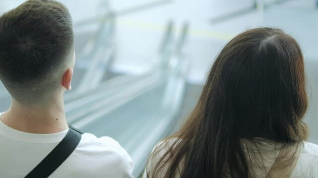 Woman with long brown hair is walking down an escalator with a man behind her. The escalator is crowded with people, and the woman is looking down at her feet. Scene is somewhat somber