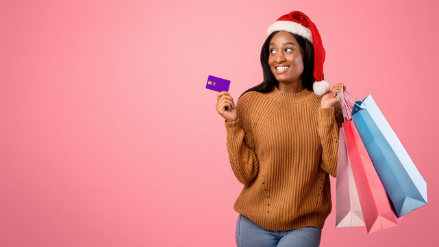 Xmas shopping concept. Pretty black woman in Santa hat holding gift bags and credit card on pink studio background, copy space. Lovely African American lady buying presents for Christmas
