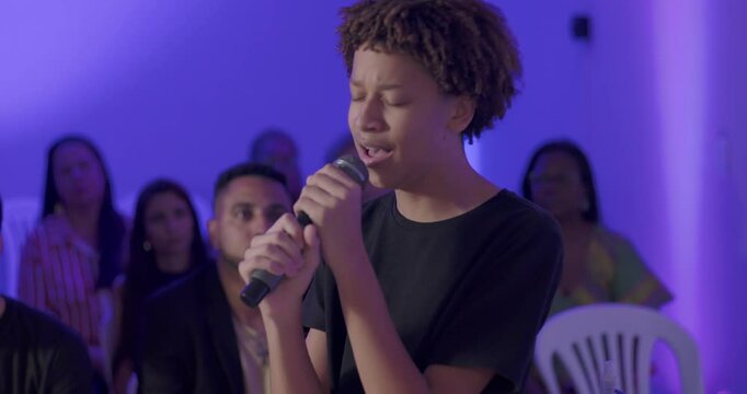 Young African American teenager singing passionately with microphone during Christian evangelical worship service, congregation in background