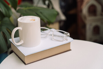 White mug with coffee foam on a closed book, next to eyeglasses and green leaves.