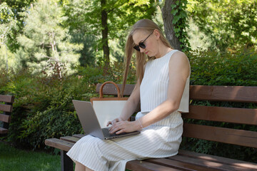 A woman is sitting on a bench and typing on a laptop. She is wearing sunglasses and a white dress