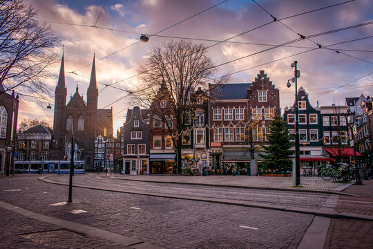 Amsterdam, Netherlands. A city square with cobblestone streets and traditional Dutch buildings adorned with holiday lights. A tram is making its way through the square along the tracks.