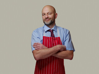 Portrait of a professional butcher in blue shirt with tie and red and white stripes apron, cream color clean background. Man in his 40s with grey short beard and bold head. Friendly face.