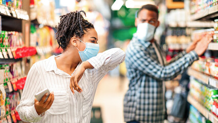 Sick African Woman In Supermarket Coughing In Elbow Wearing Mask Doing Grocery Shopping Buying Food Indoor. Shop Safe, Coronavirus Covid-19 Pandemic Outbreak, Social Distance In Public Places.