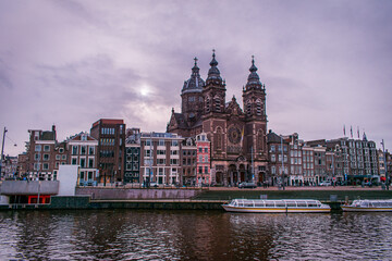 Amsterdam, Netherlands. The features boats docked along the canal. In the background, the Basilica of Saint Nicholas with multiple spires and domes. 