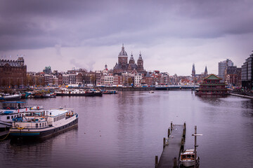 Fototapeta premium Amsterdam, Netherlands. The features boats docked along the canal. In the background, the Basilica of Saint Nicholas with multiple spires and domes. 